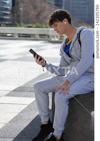 Teen male sitting on concrete seat in plaza holding phone with earbuds gray tracksuit, copy space Teen male sitting on concrete seat in plaza holding phone with earbuds gray tracksuit, copy space 142305760