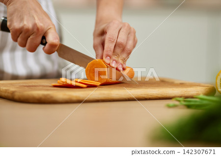 Hands slicing fresh carrot on wooden cutting board Hands slicing fresh carrot on wooden cutting board 142307671