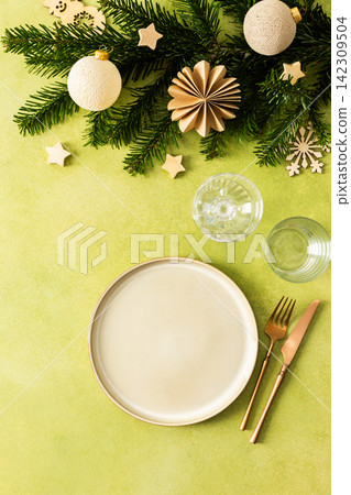A festive Christmas table setting with an empty plate and cutlery on a bright green background. Spruse branches and golden tree decorations create a warm, holiday atmosphere A festive Christmas table setting with an empty plate and cutlery on a bright green background. Spruse branches and golden tree decorations create a warm, holiday atmosphere 142309504