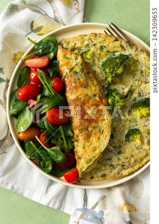 A potato omelette made with eggs and mixed greens is served on a white plate. Spinach, cherry tomatoes, and broccoli are arranged on the side for a healthy breakfast option A potato omelette made with eggs and mixed greens is served on a white plate. Spinach, cherry tomatoes, and broccoli are arranged on the side for a healthy breakfast option 142309655