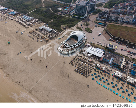 High angle wide horizontal aerial drone photograph of Scheveningen beach promenade, modern beach clubs, curved restaurant, dunes, and coastal skyline in Scheveningen, The Hague, Netherlands 142309855