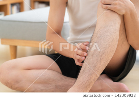 A young man removes hair from his legs at home. A young man removes hair from his legs at home. 142309928