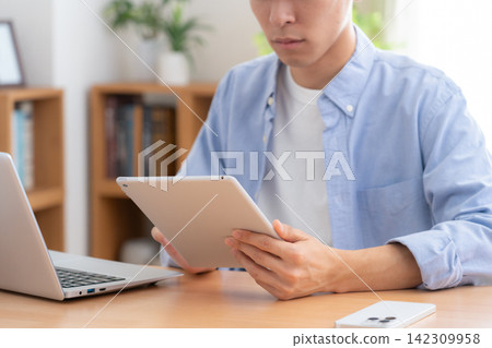 一位年輕男子在家中看著他的筆記型電腦和平板電腦。 142309958