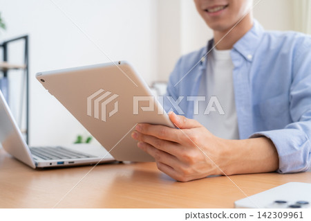 一位年輕男子在家中看著他的筆記型電腦和平板電腦。 142309961