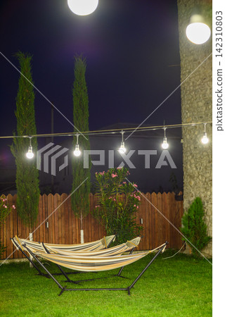 A serene nighttime outdoor scene featuring two hammocks in a well-lit private backyard with lush greenery and a wooden fence A serene nighttime outdoor scene featuring two hammocks in a well-lit private backyard with lush greenery and a wooden fence 142310803