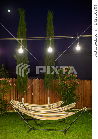 A serene nighttime backyard featuring two well-maintained hammocks on a grassy lawn illuminated by warm white lights A serene nighttime backyard featuring two well-maintained hammocks on a grassy lawn illuminated by warm white lights 142310804