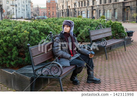 Man student sitting on bench and drinking coffee in city park. Young guy wearing fur hat resting outdoors with hot beverage. Lifestyle leisure in cold weather, urban street scene. Man student sitting on bench and drinking coffee in city park. Young guy wearing fur hat resting outdoors with hot beverage. Lifestyle leisure in cold weather, urban street scene. 142311998