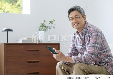 一位六十多歲的男子,面帶微笑,手持智慧型手機,直視鏡頭。 一位六十多歲的男子,面帶微笑,手持智慧型手機,直視鏡頭。 142312226