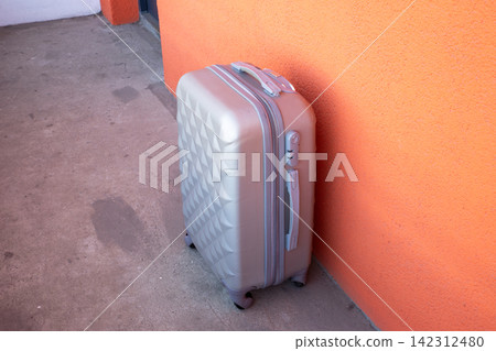 Silver suitcase standing against an orange wall. Travel luggage for vacation and business trip. Concept of tourism, summer holiday and adventure, departure for airport transport journey. Silver suitcase standing against an orange wall. Travel luggage for vacation and business trip. Concept of tourism, summer holiday and adventure, departure for airport transport journey. 142312480