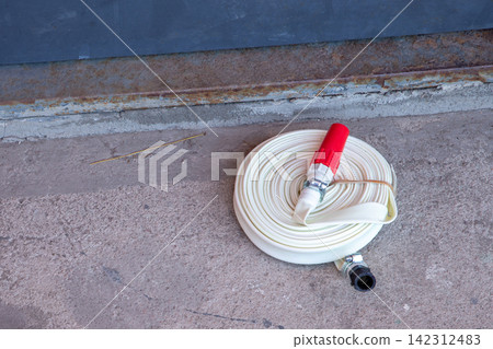 White fire hose coiled on concrete floor next to wall. Professional firefighting equipment for emergency water supply and fire safety prevention. Industrial safety and utility tool. White fire hose coiled on concrete floor next to wall. Professional firefighting equipment for emergency water supply and fire safety prevention. Industrial safety and utility tool. 142312483