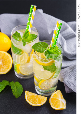 Cold homemade lemonade with fresh lemon, mint and ice in glasses on black textured stone background Cold homemade lemonade with fresh lemon, mint and ice in glasses on black textured stone background 142312860