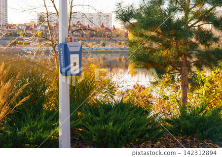 Emergency call box, panic alarm in autumn park by lake with pine tree and city view background Emergency call box, panic alarm in autumn park by lake with pine tree and city view background 142312894