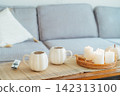 Autumn fall mood composition for hygge home decor. White pumpkin's shaped cups with hot drink, burning candles, remote controller on coffee table in living room. Movie night at home. Cozy relax time. 142313100