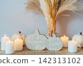 Cozy warm autumn home interior - white knitted pumpkins, wooden statuette, candles, dried flowers on the wooden console with white wall background. Scandinavian minimalist hygge home fall decor. 142313102