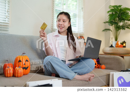 Asian woman holding credit card and using laptop on sofa for Halloween online shopping Asian woman holding credit card and using laptop on sofa for Halloween online shopping 142313382