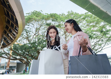 Two Asian women smiling while looking into shopping bags outdoors at mall Two Asian women smiling while looking into shopping bags outdoors at mall 142313383