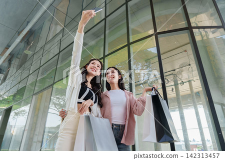 Asian women taking selfie with smartphone while carrying shopping bags outside mall Asian women taking selfie with smartphone while carrying shopping bags outside mall 142313457