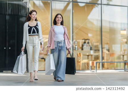 Two smiling Asian women walking outside glass storefront carrying shopping bags Two smiling Asian women walking outside glass storefront carrying shopping bags 142313492