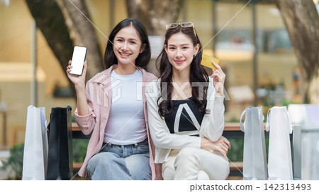 Asian women holding smartphone and credit card while sitting with shopping bags outdoors Asian women holding smartphone and credit card while sitting with shopping bags outdoors 142313493