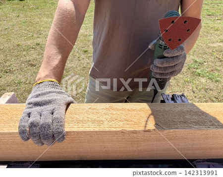 A carpenter holds a sander and rests his hand on a wooden beam outdoors. A carpenter holds a sander and rests his hand on a wooden beam outdoors. 142313991
