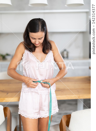 Adult woman measuring body with tape measure in kitchen Adult woman measuring body with tape measure in kitchen 142314377