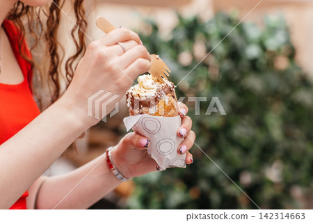 Close up of woman hands holding sweet pastry with cream and chocolate topping outdoors Close up of woman hands holding sweet pastry with cream and chocolate topping outdoors 142314663