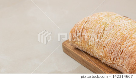 Freshly baked ciabatta bread loaf resting on a wooden cutting board against a light textured background, showcasing its crusty exterior and unique shape. space for text Freshly baked ciabatta bread loaf resting on a wooden cutting board against a light textured background, showcasing its crusty exterior and unique shape. space for text 142314667