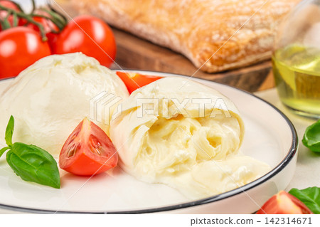 Fresh burrata cheese served on a white plate with cherry tomatoes, basil leaves, and a slice of bread, drizzled with olive oil in a rustic kitchen setting Fresh burrata cheese served on a white plate with cherry tomatoes, basil leaves, and a slice of bread, drizzled with olive oil in a rustic kitchen setting 142314671