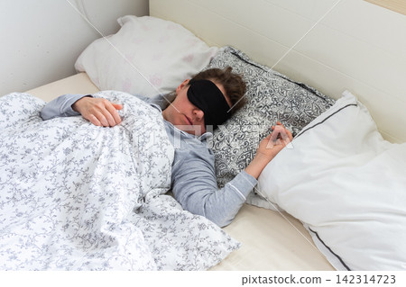 A 50-year-old woman is comfortably sleeping in her bed with a sleep mask on. She is wrapped in a soft blanket, resting peacefully in a serene bedroom setting A 50-year-old woman is comfortably sleeping in her bed with a sleep mask on. She is wrapped in a soft blanket, resting peacefully in a serene bedroom setting 142314723