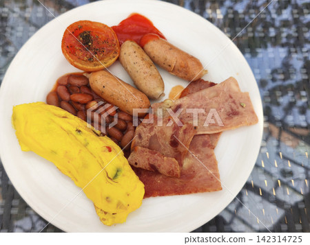 Full breakfast plate featuring grilled sausages, crispy bacon, ham, rolled omelette, baked beans, and cherry tomatoes served on a white dish. Full breakfast plate featuring grilled sausages, crispy bacon, ham, rolled omelette, baked beans, and cherry tomatoes served on a white dish. 142314725