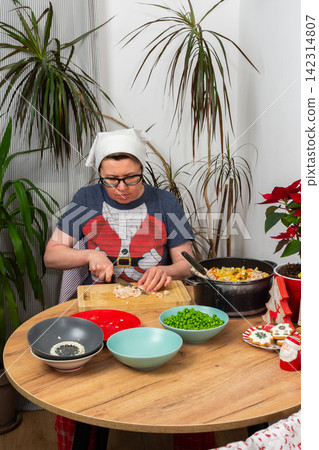 A woman is in her kitchen preparing Olivier salad, Russian salad, for a Christmas and New Year celebration. She is slicing boiled meat on a cutting board with several bowls of ingredients on the table A woman is in her kitchen preparing Olivier salad, Russian salad, for a Christmas and New Year celebration. She is slicing boiled meat on a cutting board with several bowls of ingredients on the table 142314807