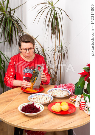 A woman grates carrots in the kitchen for making herring under fur coat salad, the traditional Ukrainian or Russian Christmas salad Shuba. Nearby are bowls with beets, potatoes, and a festive plant 142314809