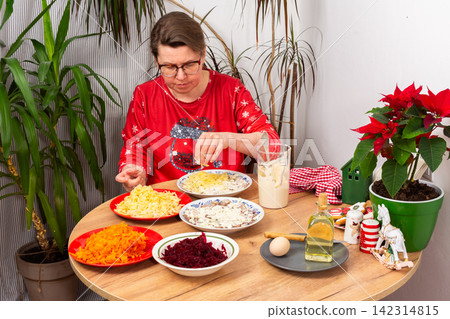 A woman spreads mayonnaise on layers of Herring Under Fur Coat salad at her kitchen table, Christmas salad Shuba She uses ingredients like herring, eggs, beets, and carrots while celebrating Christmas A woman spreads mayonnaise on layers of Herring Under Fur Coat salad at her kitchen table, Christmas salad Shuba She uses ingredients like herring, eggs, beets, and carrots while celebrating Christmas 142314815