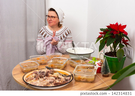 A woman stands in her cozy kitchen preparing kholodets, a traditional Ukrainian dish made with jellied meat. She focuses on the table filled with containers ready for Christmas dinner 142314831