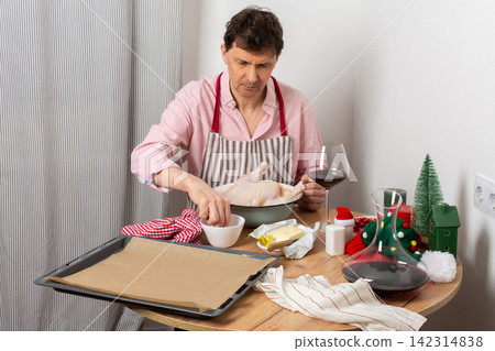 A man is preparing for a pre-Christmas dinner by baking chicken and potatoes. He is focused on his task while surrounded by festive decorations and a glass of wine A man is preparing for a pre-Christmas dinner by baking chicken and potatoes. He is focused on his task while surrounded by festive decorations and a glass of wine 142314838