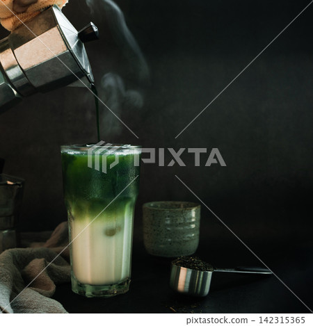 Poured Matcha green tea with Italian Moka pot pouring into a cup with iced of milk and powdered green tea on spoon on black background. copy space for your text Poured Matcha green tea with Italian Moka pot pouring into a cup with iced of milk and powdered green tea on spoon on black background. copy space for your text 142315365