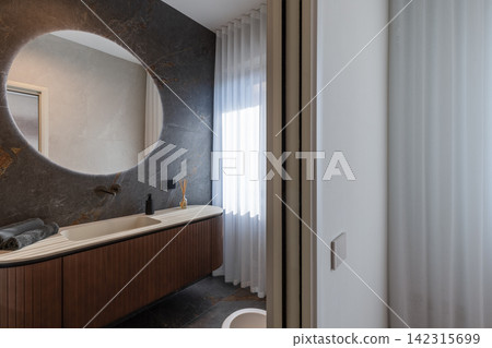 Bathroom vanity with a backlit round mirror against textured panels and pale surfaces Bathroom vanity with a backlit round mirror against textured panels and pale surfaces 142315699