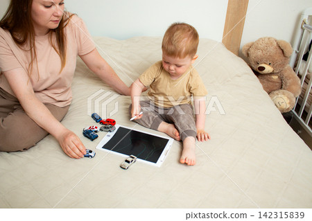Mother and two year old son drawing on kids tablet with stylus. Parent child bonding through creative play. Fine motor skills developing while making digital art together Mother and two year old son drawing on kids tablet with stylus. Parent child bonding through creative play. Fine motor skills developing while making digital art together 142315839