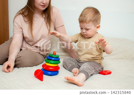 Toddler placing bright rings on pyramid pole with mom's guidance. Learning shapes, sizes and sequence. Early education through hands on play. Bonding time Toddler placing bright rings on pyramid pole with mom's guidance. Learning shapes, sizes and sequence. Early education through hands on play. Bonding time 142315840