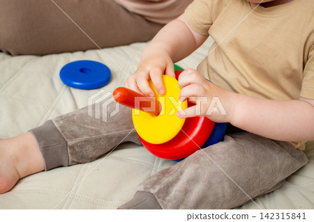 Mother helping child build pyramid on cozy bed. Educational play with shapes and colors. Parent interaction into child game. Family bonding concept Mother helping child build pyramid on cozy bed. Educational play with shapes and colors. Parent interaction into child game. Family bonding concept 142315841