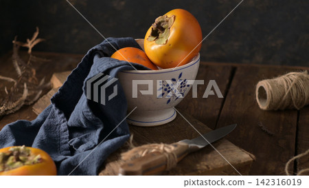 Fresh ripe persimmons in a patterned bowl on a wooden board with a knife and cloth Fresh ripe persimmons in a patterned bowl on a wooden board with a knife and cloth 142316019