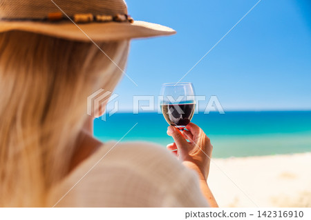 Woman in straw hat holding glass of red wine while enjoying turquoise sea view and relaxing on sunny beach vacation Woman in straw hat holding glass of red wine while enjoying turquoise sea view and relaxing on sunny beach vacation 142316910