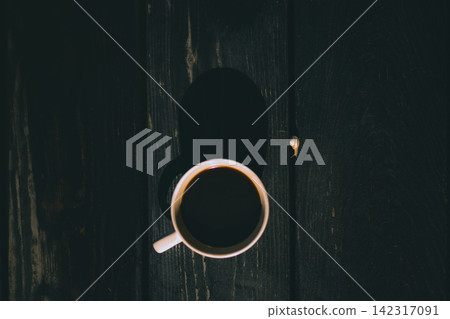Top view of a cup of black coffee on a dark wooden table, with strong shadows and minimalist composition. The moody still life evokes quiet mornings, solitude, simplicity and slow living. 142317091