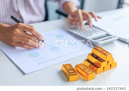 Selective focus on gold bars with background of hands using calculator for gold trading analysis. Concept for wealth management, asset protection, and financial planning. Selective focus on gold bars with background of hands using calculator for gold trading analysis. Concept for wealth management, asset protection, and financial planning. 142317441