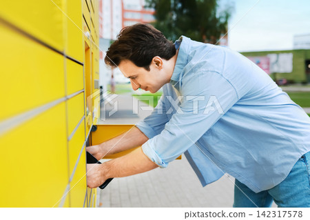 man picking up an online shopping package from a yellow parcel locker. A convenient contactless delivery service, ecommerce logistics, and self-service terminal. man picking up an online shopping package from a yellow parcel locker. A convenient contactless delivery service, ecommerce logistics, and self-service terminal. 142317578