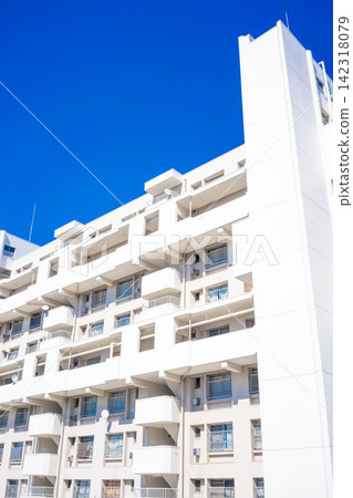 The white exterior of the apartment complex stands out against the dazzling blue sky, showcasing the modern architectural beauty of the housing complex. The white exterior of the apartment complex stands out against the dazzling blue sky, showcasing the modern architectural beauty of the housing complex. 142318079