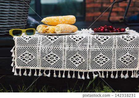 Outdoor table covered with a white crocheted lace tablecloth, with yellow sunglasses, pastries and red berries arranged on top. Rustic picnic setting evokes summer, leisure, traditional textile decor. Outdoor table covered with a white crocheted lace tablecloth, with yellow sunglasses, pastries and red berries arranged on top. Rustic picnic setting evokes summer, leisure, traditional textile decor. 142318231