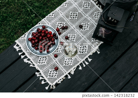 Top view of a rustic outdoor picnic with red berries, a glass of lemonade and a white crocheted lace runner on a dark wooden table. Summer setting evokes handmade decor, leisure countryside lifestyle. 142318232