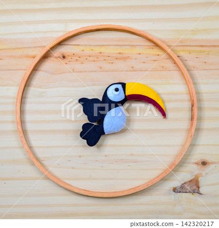 Handmade felt toucan bird decoration arranged inside a circular wooden ring on a light wooden background Handmade felt toucan bird decoration arranged inside a circular wooden ring on a light wooden background 142320217
