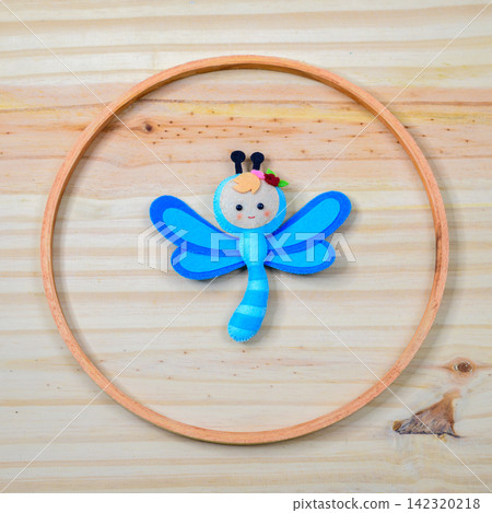 Handmade felt dragonfly decor arranged inside a circular wooden ring on a light wooden background Handmade felt dragonfly decor arranged inside a circular wooden ring on a light wooden background 142320218